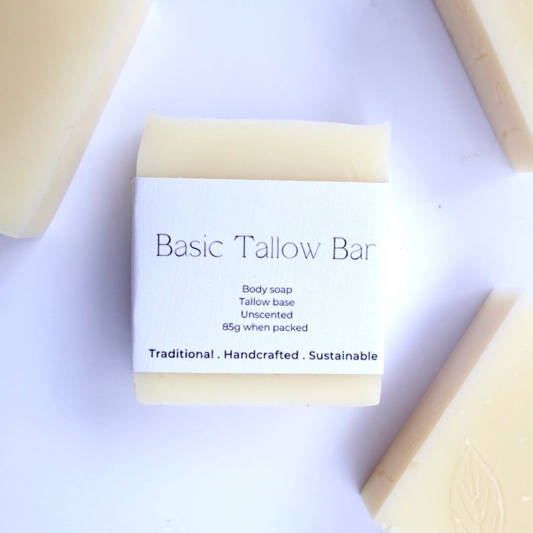 A rectangular bar of Basic Tallow Bar body soap with a white paper label, surrounded by other soap bars on a white background.