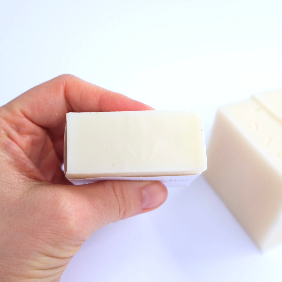 A hand holding a rectangular bar of creamy tallow off-white soap against a plain white background, with another soap bar blurred in the background.