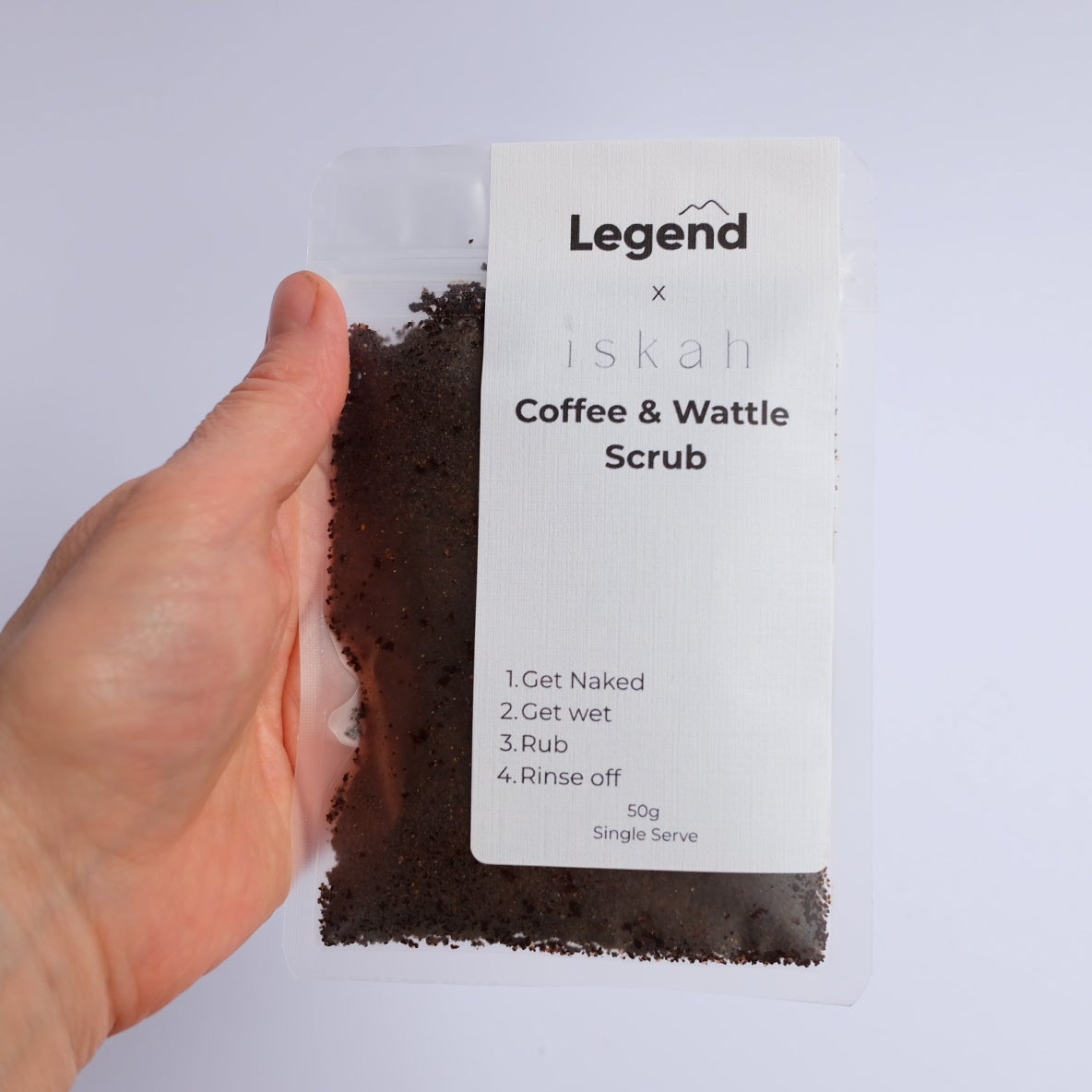 A hand holds a clear plastic pouch of 'Legend x iskah Coffee & Wattle Scrub' against a plain light gray background. The package contains dark brown scrub and features a white label with black text.